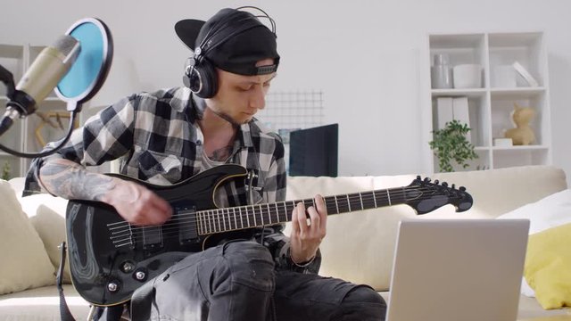 Tilt up of young musician sitting on sofa in the living room, playing electric guitar and using laptop while recording song in home studio