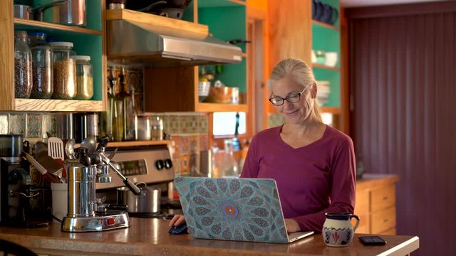 Camera moving right as casually dressed mature woman smiles and works on laptop computer and drinks tea in kitchen.