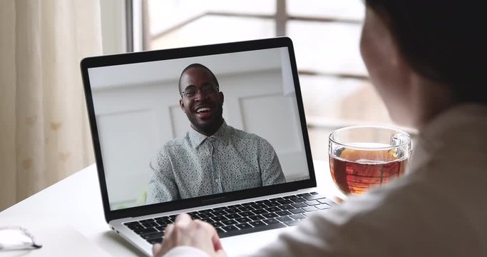 Happy african man teacher, friend, remote worker talks with caucasian woman student study work from home office, makes conference video call virtual chat on laptop screen. Over shoulder closeup view.