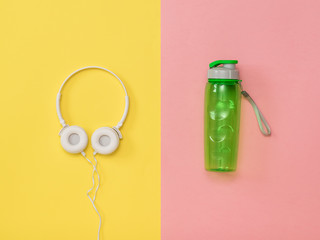 White headphones and a green sports water bottle on a yellow and pink background. Flat lay.