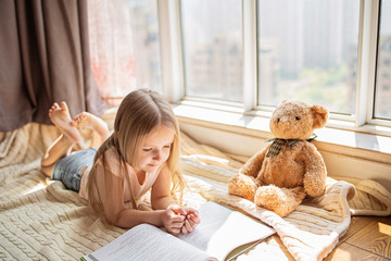 Cute little caucasian girl in casual clothes reading a book with stuffed teddy bear toy and smiling while lying on a floor near window in the room. Stay at home during coronavirus covid-19 pandemic