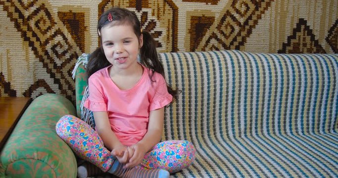 Smiling and happy cute girl of five years old with brown hair and blue eyes in pink T-shirt sitting at home on sofa covered with bright blanket looking to camera and waving her hand