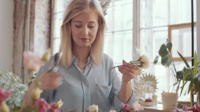 Tilt up shot of beautiful blonde female florist cutting flower stem with pruning shears while making composition in hat box