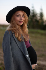 Young pregnant caucasian woman with long fair hair in black hat and grey coat outside on warm day. Smiles and looks at camera. Side view. Nature backsides. Expectation of new life concept