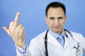 Middle-aged angry doctor in white medical coat and stethoscope showing middle finger doing fuck you, provocation and rude attitude, over blue background