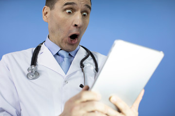 Portrait of a shocked male doctor with stethoscope dressed in uniform looking at laptop tablet while standing isolated over blue background.