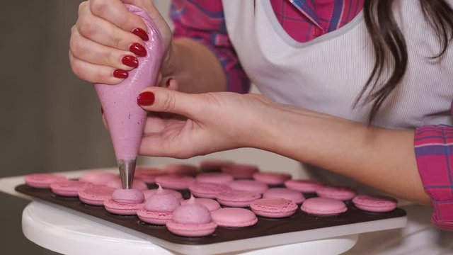 A smiling woman baker is making macarons with a pastry bag. The woman is putting filling for the tasty home-made macarons.