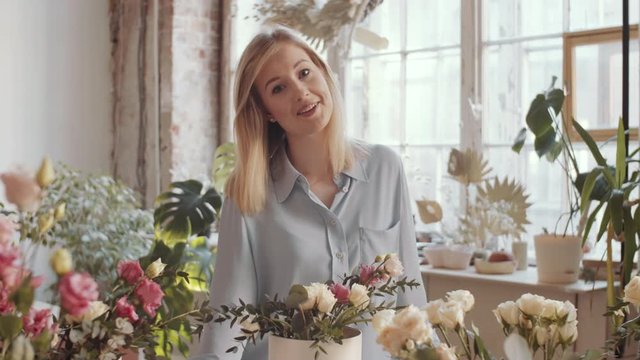 Arc shot of beautiful blonde female florist arranging roses composition in hatbox and then looking at camera and happily smiling while posing at workplace in flower shop