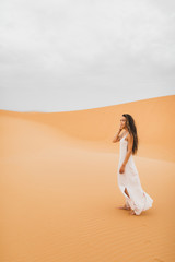 Fototapeta na wymiar Pretty portrait of young beautiful woman in sand dunes of moroccan Sahara desert. Brunette with long hair, eastern appearance. Bride in white silk dress.
