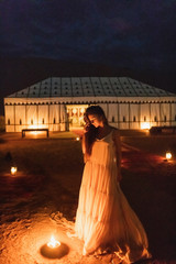 Woman in white dress at night near burning candle. Romantic evening in glamping desert camp in Sahara, Morocco. Unusual destination for wedding. - 337586344