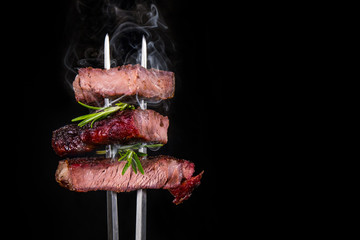 Grilled ribeye beef steak with rosemary on a black background. Beef steak on a fork. Hot pieces of...