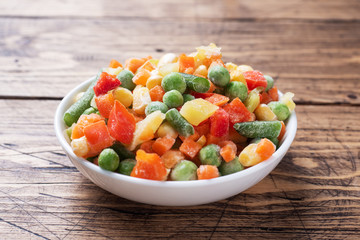 Chopped frozen vegetables in a plate on a wooden background. Corn peas pepper carrots. copy space.