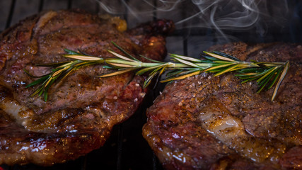 Ribeye beef steak on a grill with rosemary branch. Hot two pieces of cow meat with smoke.