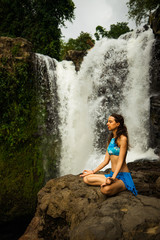 Young Caucasian woman sitting on the rock, meditating, practicing yoga at waterfall. Gyan mudra. Tegenungan waterfall in Ubud, Bali, Indonesia.