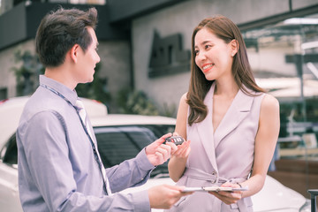 Asian officer male staff stands and holding rental agreement document in front with customer presenting new cars at showroom. Young Asian  woman buyer signing contract for new car. Car rental concept.