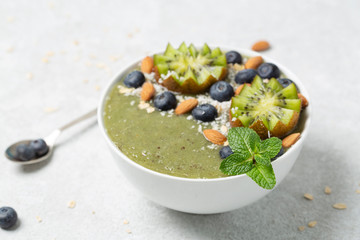 Breakfast green smoothie bowl topped with kiwi, blueberry, almond and coconut flakes on light grey background. 