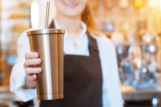 Closeup female hands are holding stainless metal reusable tumbler cup mug with drinking straw. Barista woman  prepared, brewed coffee using professional machine in cafe, restaurant. Take away, to go.