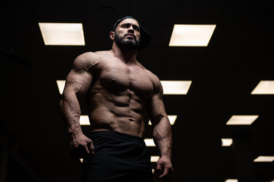 muscular aggressive young male with beard wearing cap in dark sport gym with yellow lamps light