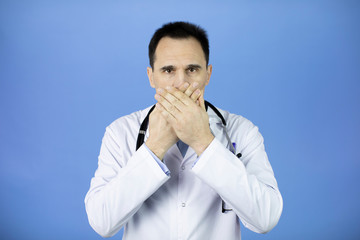 Portrait of a Caucasian adult doctor in a white coat with a stethoscope over his neck on a blue background. He covers his mouth with his palms. Confidential information. Prohibition of disclosure.