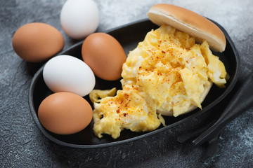Burger bun with american style scrambled eggs in a cast-iron serving pan, horizontal shot on a dark grey stone background