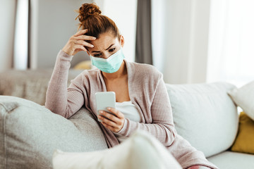 Worried woman with face mask texting on cell phone at home.