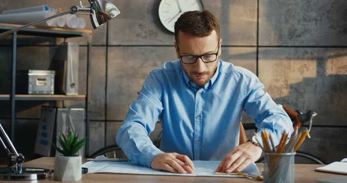 Stylish serious male professional drawing with pencils plan while sitting at office desk. Handsome busy Caucasian businessman with glasses drafting scheme at workplace indoor. Work concept