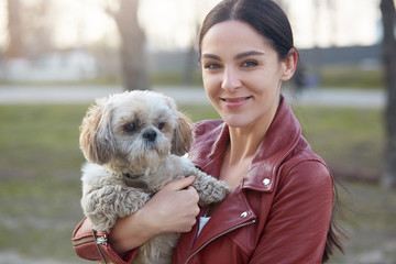 Image of attractive charismatic brunette looking directly at camera, having pleasant facial expression, holding her Maltese dog, being fond of animals, enjoying spring time. People and pets concept.