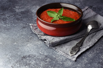 Traditional Spanish cold summer soup gazpacho. Tomato soup decorated with leaf of basil. Textured stone background with place for text. Soup in bowl with spoon on grey towel.