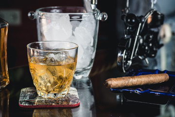 Glass of whiskey and cigar next to an ice bucket on a glass table