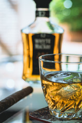 Glass of whiskey and cigar next to a bottle on glass table
