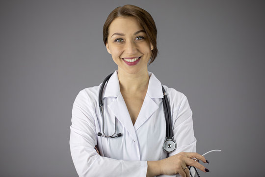 Portrait of an attractive sexy doctor woman in a white medical coat with a stethoscope on her neck. Hands are folded on the chest. Red painted lips, a snow-white smile. Grey background. Close-up