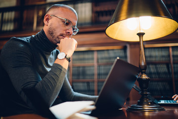 Adult 30 years old male student spending time in university library for learning, intelligent man in optical bifocal spectacles searching scientific information via app on modern laptop computer