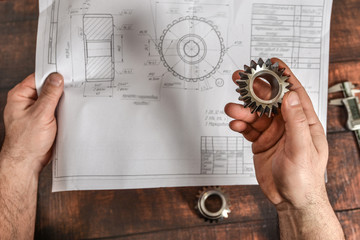 Fototapeta na wymiar The working engineer holds a bevel gear in his hand against the background of a technical drawing.