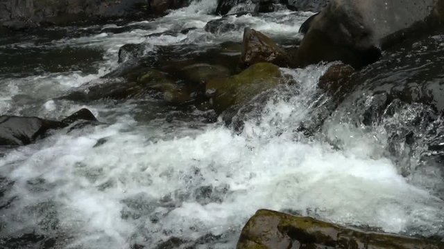 Stream, a body of water flowing through rocky creek parted of big waterfall, forming a white water.  Close up.