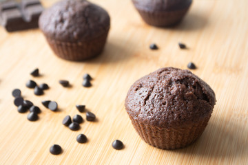 Chocolate muffins, chocolate chips, chocolate bar, a cup of coffee, teapot on a wooden surface with a warm scarf, Christmas lights bokeh and holly in the background