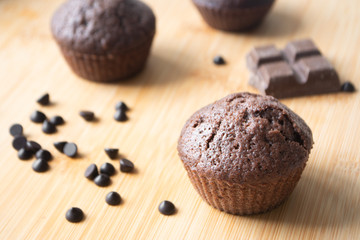 Chocolate muffins, chocolate chips, chocolate bar, a cup of coffee, teapot on a wooden surface with a warm scarf, Christmas lights bokeh and holly in the background