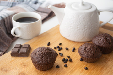 Chocolate muffins, chocolate chips, chocolate bar, a cup of coffee, teapot on a wooden surface with a warm scarf, Christmas lights bokeh and holly in the background