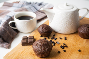Chocolate muffins, chocolate chips, chocolate bar, a cup of coffee, teapot on a wooden surface with a warm scarf, Christmas lights bokeh and holly in the background