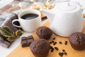 Chocolate muffins, chocolate chips, chocolate bar, a cup of coffee, teapot on a wooden surface with a warm scarf, Christmas lights bokeh and holly in the background