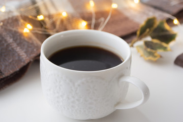 A cup of coffe, chocolate and a pot with warm scarf holly and christmas lights bokeh in the background for Scandinavian hygge atmosphere