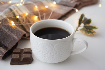 A cup of coffe, chocolate and a pot with warm scarf holly and christmas lights bokeh in the background for Scandinavian hygge atmosphere