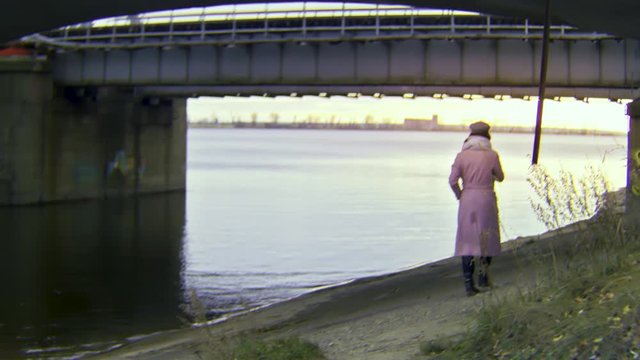 Rear view of a lonely beautiful woman in warm autumn clothes walking under the bridge near the river. Stock footage. Retro camera effect, romantic girl walking in an industrial area.