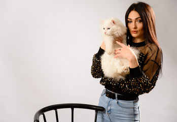 Photo of a cute caucasian brunette girl with good makeup in a shiny blouse with a white cat in her arms. Made in the studio on a white background.