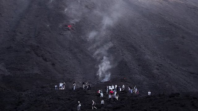 A big group of tourists stand dangerously close to a smoking lava flow as a large molten rock rolls down the black volcano towards them