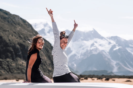 Two happy young girls who are travel lovers enjoy the moment side by side on a roadtrip. They are on top of the van laughing in front of the mountains and getting lost in the nature.Travel concept.