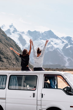 Two adventurous female travelers raise their hands in front of Mount Cook lookout. They pull over to the side and sit back on top of the van to enjoy the moment together. Copyspace. Travel concept