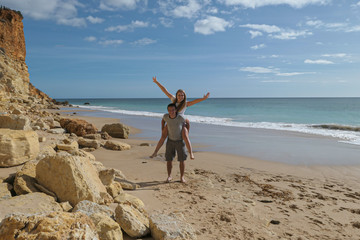 A couple near the blue sea and big rude rocs. Yellow sand on the beach.
