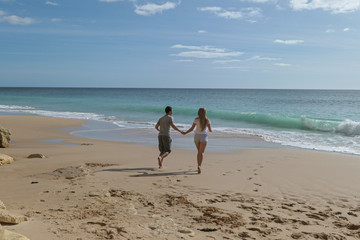 A couple near the blue sea and big rude rocs. Yellow sand on the beach.