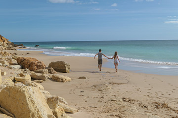 A couple near the blue sea and big rude rocs. Yellow sand on the beach.