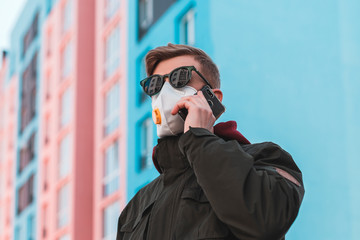 A man in glasses and a protective mask respirator talking on the phone in an empty street during a virus outbreak. A man in a protective mask calls on the phone.